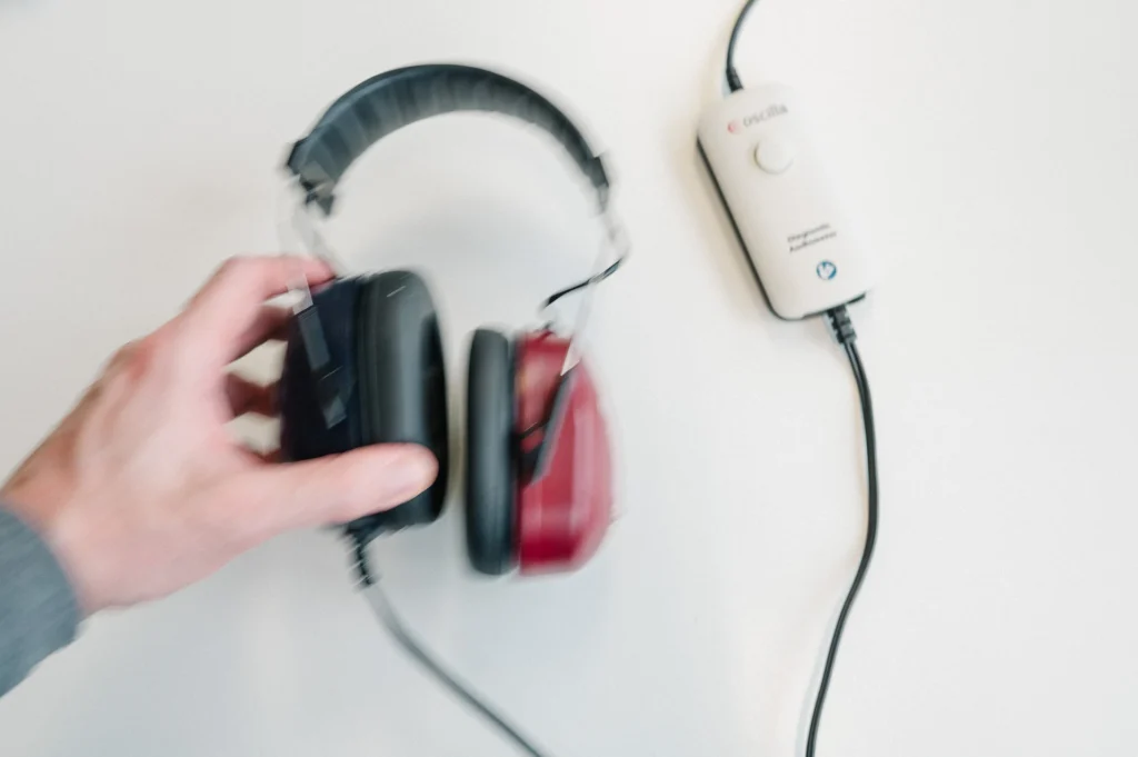 Hand hält einen Audiometrie-Kopfhörer neben einem Audiometer-Gerät der Marke Oscilla